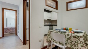 Een eetgedeelte met een tafel voor vier personen, een keuken op de achtergrond en een gang die naar een slaapkamer leidt.