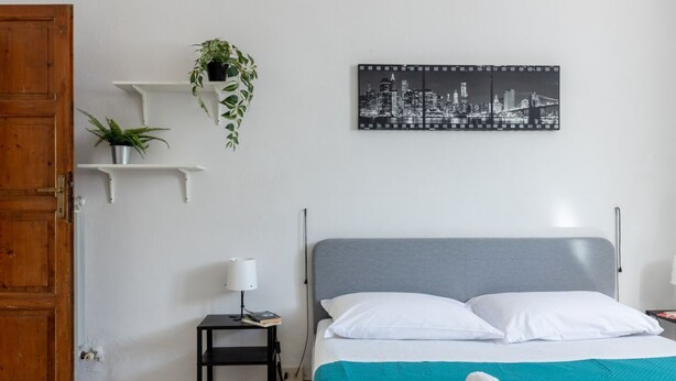A bedroom with a bed, a nightstand, a lamp, a cityscape picture, and two shelves with plants. - GuestHost - Spacious 50 sqm apartment, ideal for 4 people, located on the ground floor of a building WITHOUT lift (NOT suitable for disabled people). The apartment is located a 9-minute walk from Pisa-Galileo Galilei airport and 6 minutes by public trans (Pisa)