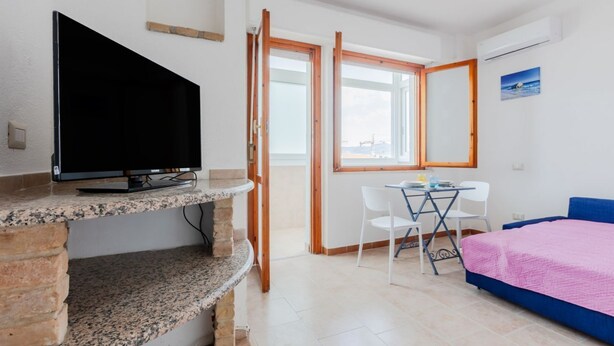 Un dormitorio con televisión, cama, escritorio y sillas, y una ventana.