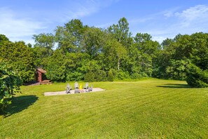 A well-maintained grassy lawn with a fire pit and chairs, surrounded by dense trees and a small wooden structure.