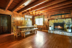 A wooden cabin interior with a stone fireplace, wooden dining table, and benches.
