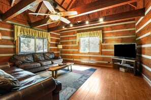 L’intérieur d’une cabane en rondins, avec un canapé en cuir, un téléviseur et un ventilateur de plafond.