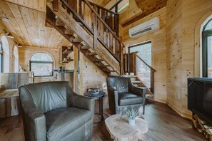 Interior de una cabaña de madera con una escalera, sillones de cuero, una mesita y un televisor.