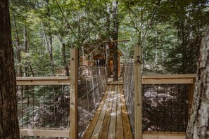 A wooden suspension bridge in a forest with a wooden cabin at the end.