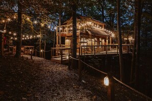 A wooden cabin with a deck, surrounded by trees and string lights.