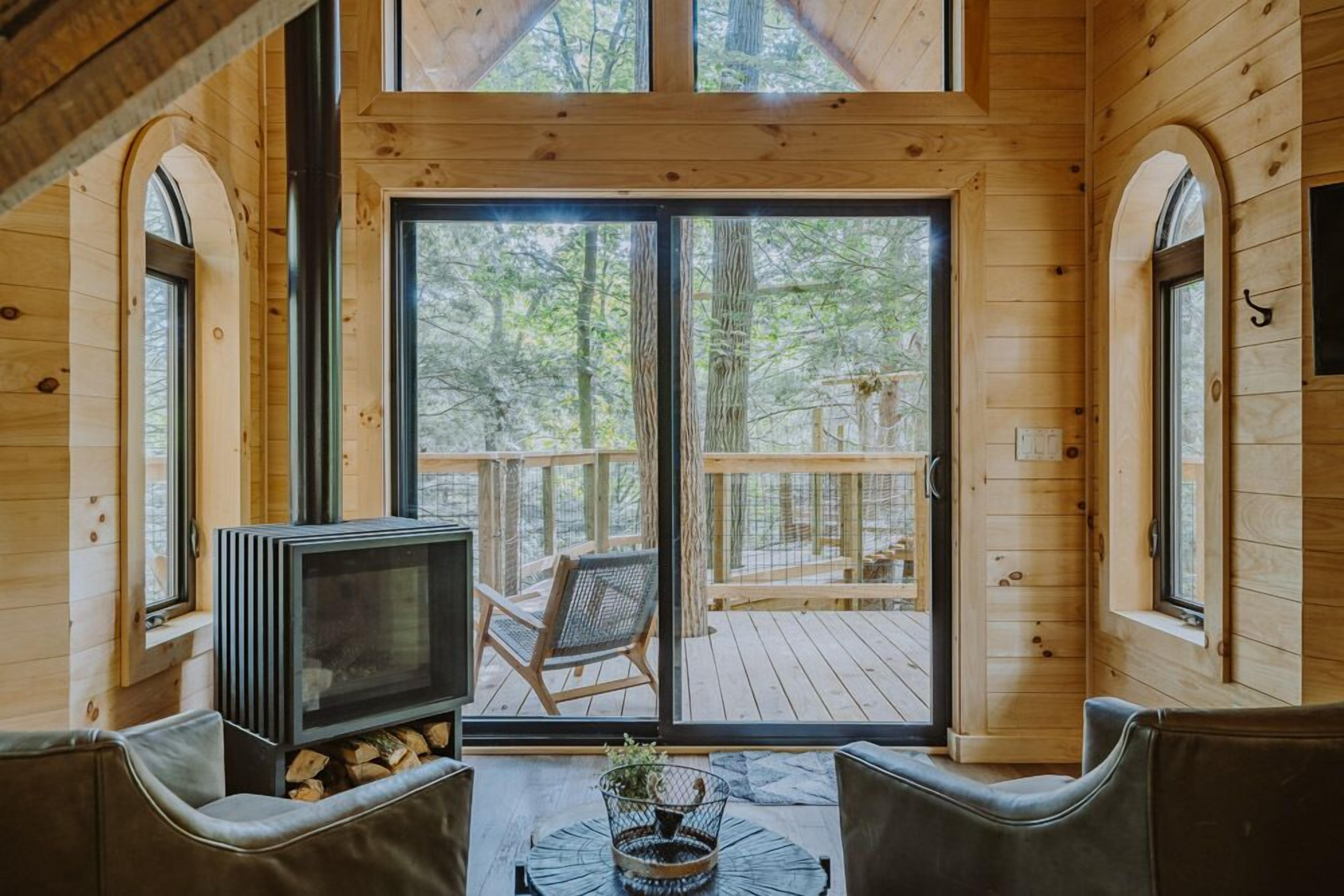 Interior de una cabaña de madera con chimenea, zona de estar y grandes puertas correderas de vidrio que dan a una terraza.