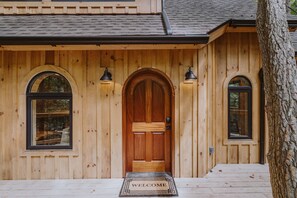A wooden house with a large arched front door, two arched windows on either side, and a welcome mat at the entrance.