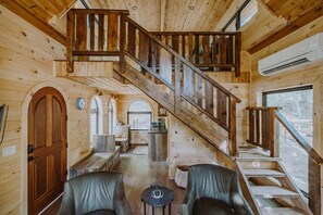 Interior de una cabaña de madera con una escalera central, un comedor y una sala de estar con sillones de cuero.