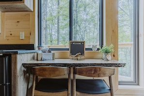 Un comptoir de cuisine en bois, accompagné de deux chaises, d’une pancarte « BIENVENUE », d’une horloge et d’une petite plante.