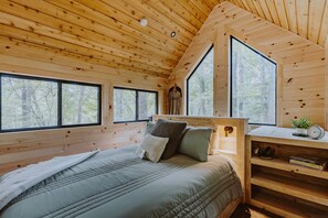 Ein Schlafzimmer in einer Holzhütte mit einem Bett, Kissen, einem Nachttisch und einem Fenster mit Blick auf Bäume.