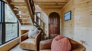 Interior de una cabaña de madera con una escalera, sofás de cuero y una puerta de madera.