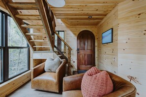 Interior de una cabaña de madera con una escalera, sofás de cuero y una puerta de madera.