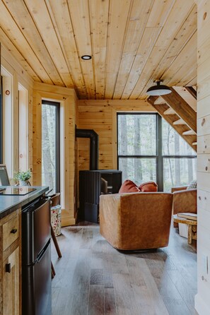 A cozy cabin interior with wooden walls, a fireplace, a dining area, and a large window offering a view of the forest.