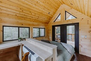A wooden cabin interior with a loft bedroom, a dining area, and large windows.