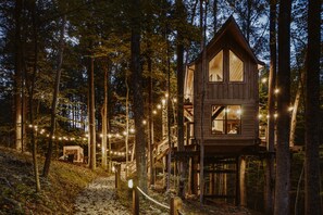 Eine hölzerne Blockhütte auf Stelzen in einem Wald, umgeben von Bäumen und beleuchtet mit Lichterketten.