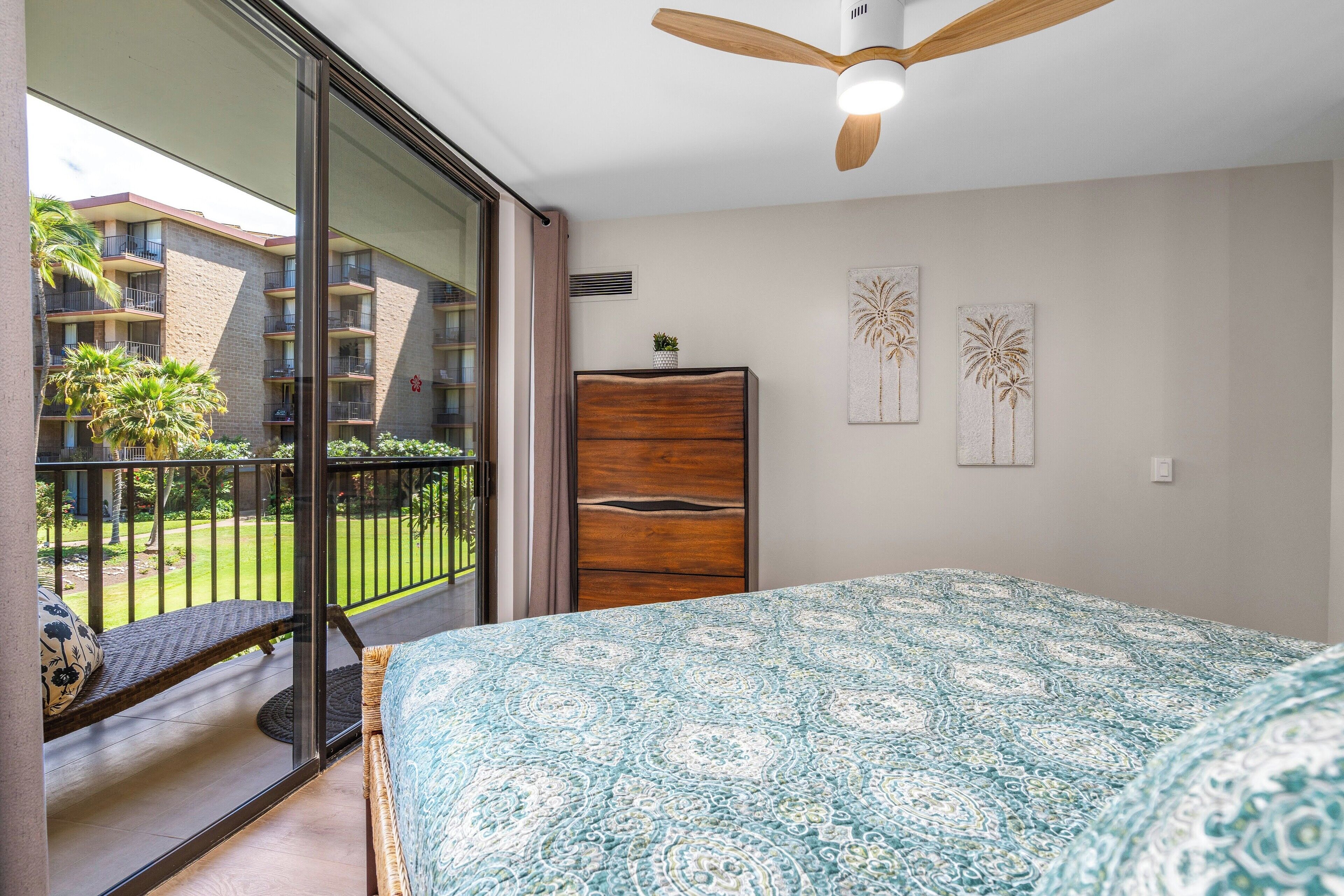 A bedroom with a large bed, a wooden wardrobe, a ceiling fan, and a balcony with a view of a building and greenery.