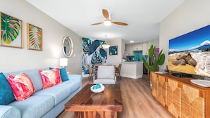 A modern living room with a sofa, coffee table, and a TV on a wooden cabinet. There is a dining area with a table and chairs, and a kitchen in the background. - Hip Hawaiian Oceanfront Vistas (Kihei)