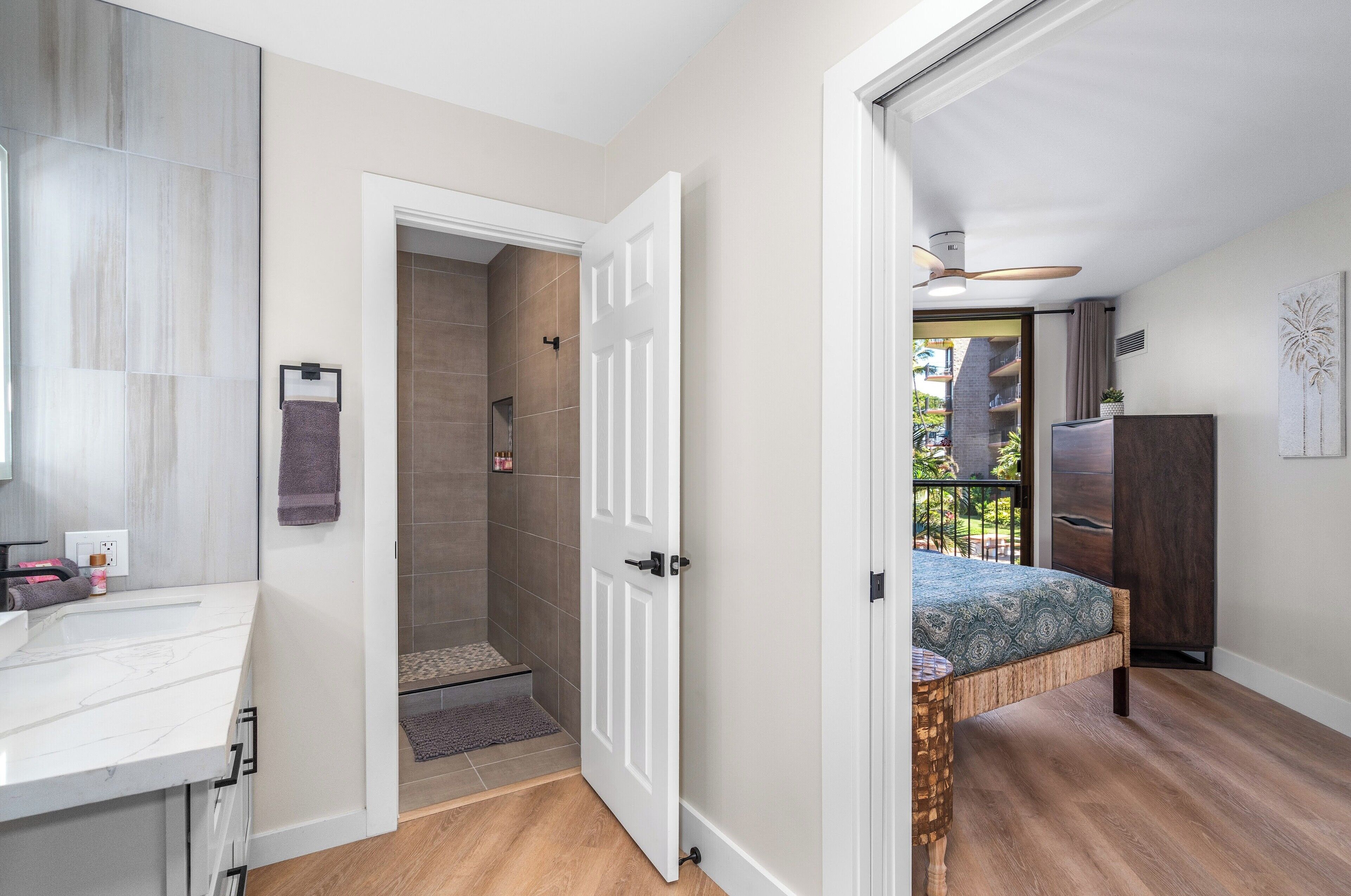 A modern bathroom with a white vanity, a shower, and a door leading to a bedroom with a bed and a balcony.