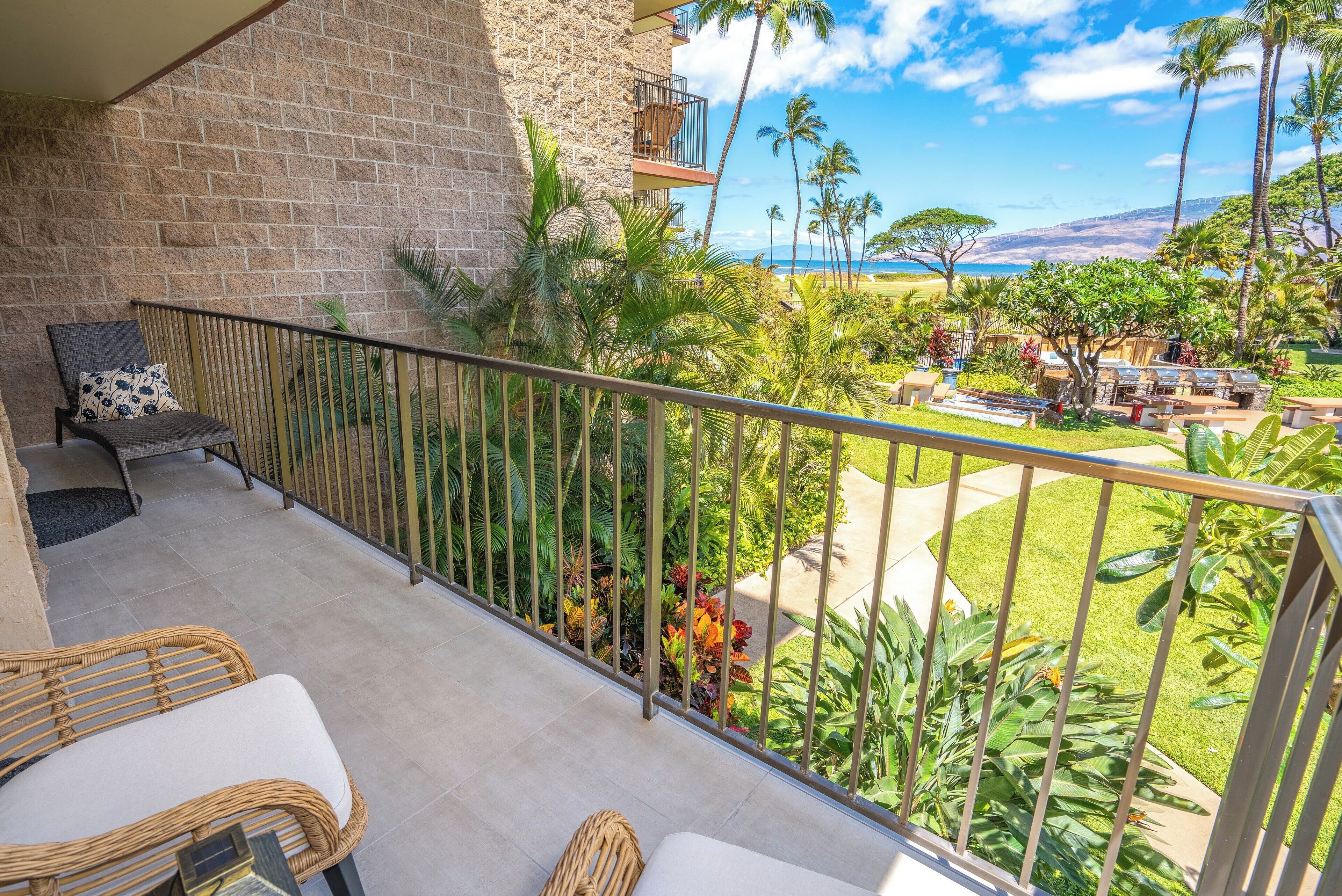 A balcony with wicker furniture, a stone wall, and a view of a tropical garden and beach.