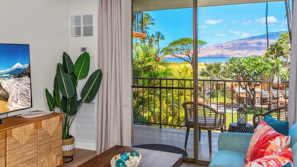 A living room with a large TV, a wooden cabinet, a plant, and a view of a balcony with outdoor furniture. - Hip Hawaiian Oceanfront Vistas (Kihei)