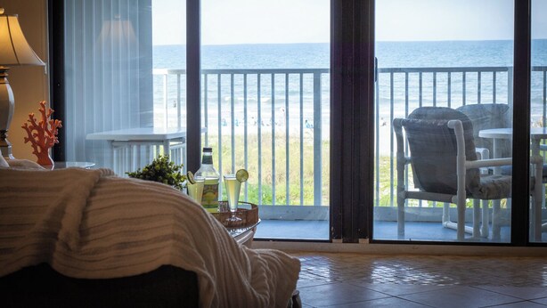 A room with a bed, a table with drinks, and a view of the ocean through large windows.