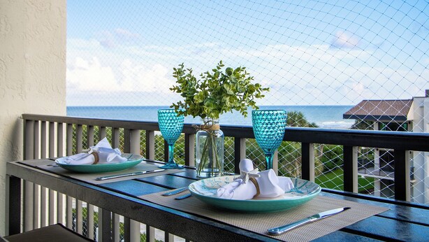 A dining table set with plates, napkins, and cutlery, a vase with greenery, and two blue glassware on a balcony overlooking the sea. - Beachside Bliss | Ocean View Condo w/ Heated Pool, Spa & Direct Beach Access 🌴 (Cape Canaveral)