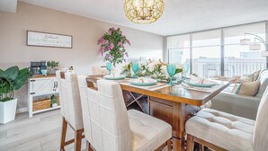 A modern dining area with a wooden table set for four, white upholstered chairs, a hanging light fixture, and a view of the city through large windows. - Beachside Bliss | Ocean View Condo w/ Heated Pool, Spa & Direct Beach Access 🌴 (Cape Canaveral)