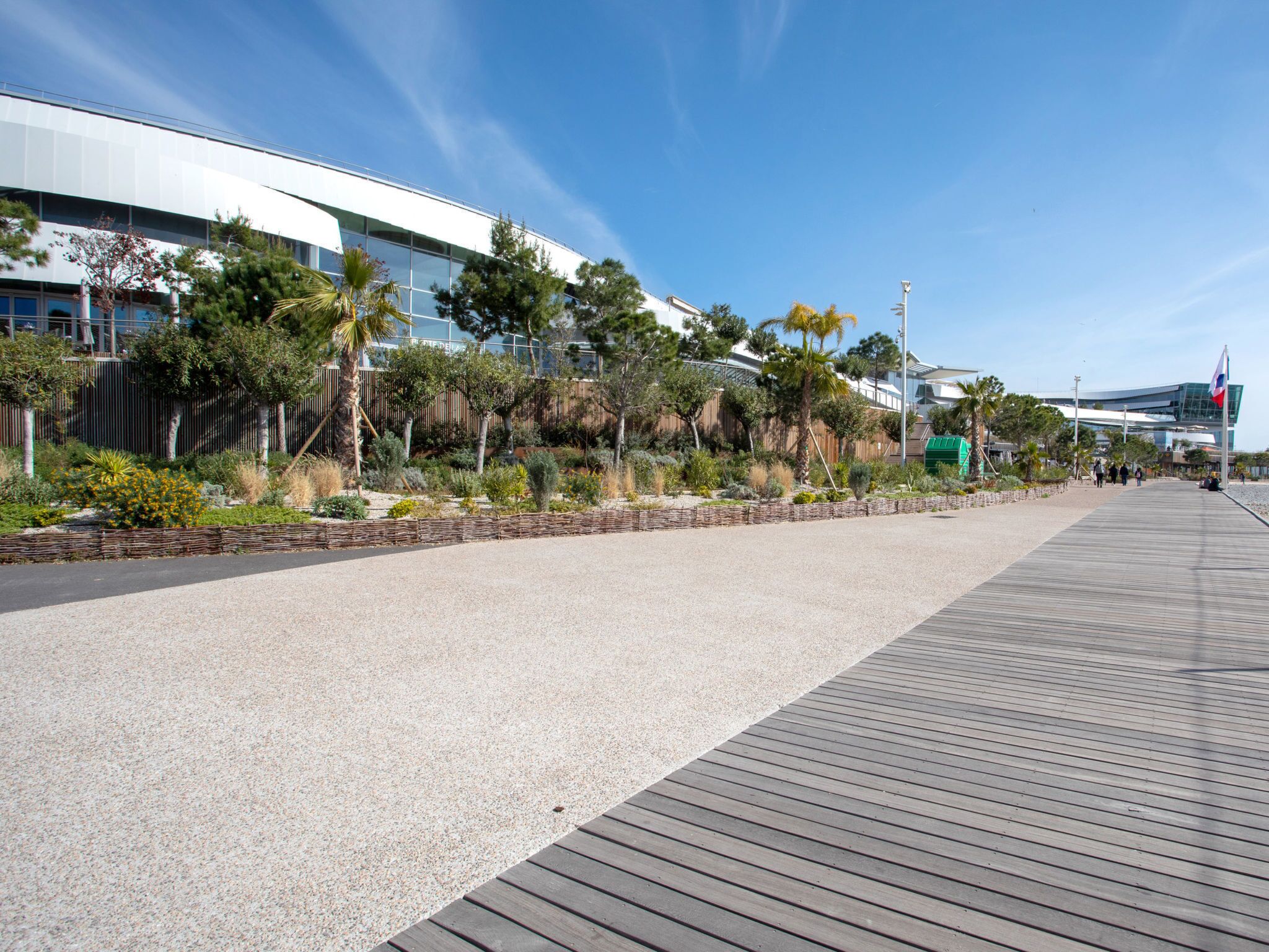 A modern building with a glass facade, a wooden boardwalk, and landscaped areas with various plants.