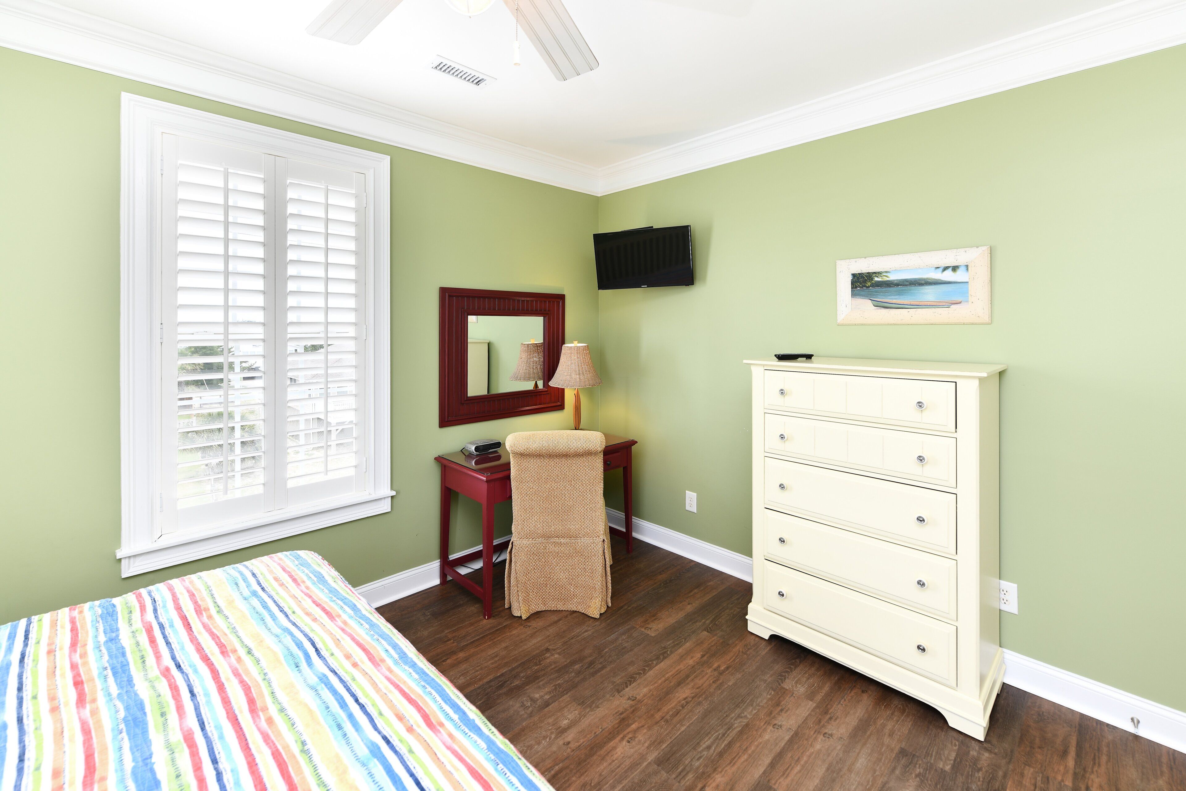 A bedroom with a bed, a desk, a chair, a dresser, a TV, and a window with blinds.