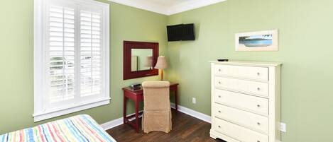 A bedroom with a bed, a desk, a chair, a dresser, a TV, and a window with blinds.
