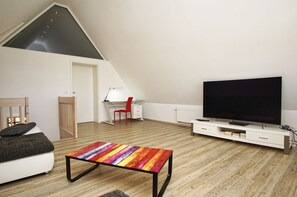 Et moderne loftværelse med skråvægge, et stort fjernsyn, et skrivebord med stol og et farverigt stribet bord.