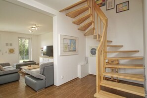 A modern living room with a wooden staircase, grey sofas, a flat-screen TV, and a large window.