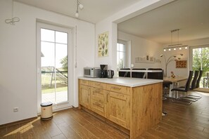 A modern kitchen with wooden cabinets, a built-in microwave, and a coffee maker. There is a dining area with a table and chairs, and a large window providing natural light.