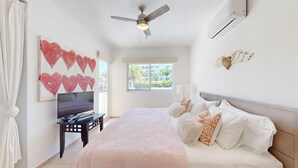 Une chambre à coucher comprenant un lit, un téléviseur, un ventilateur de plafond et une fenêtre donnant sur des immeubles.