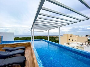Ein Infinity-Pool mit einer Holzterrasse und Liegestühlen, umgeben von einer weißen Pergola, mit Blick auf eine Stadtlandschaft und den Ozean.