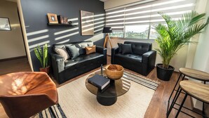Una sala de estar moderna con un sofá de cuero negro, una mesa de centro de vidrio y un cuenco de madera. Hay dos plantas en macetas, una lámpara de pie y vistas a la ciudad a través de amplios ventanales.