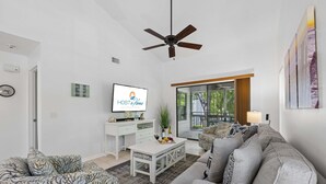 Una sala de estar moderna con televisión, ventilador de techo, sofá, mesa de centro y vistas al exterior.