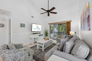 Una sala de estar moderna con televisión, ventilador de techo, sofá, mesa de centro y vistas al exterior.