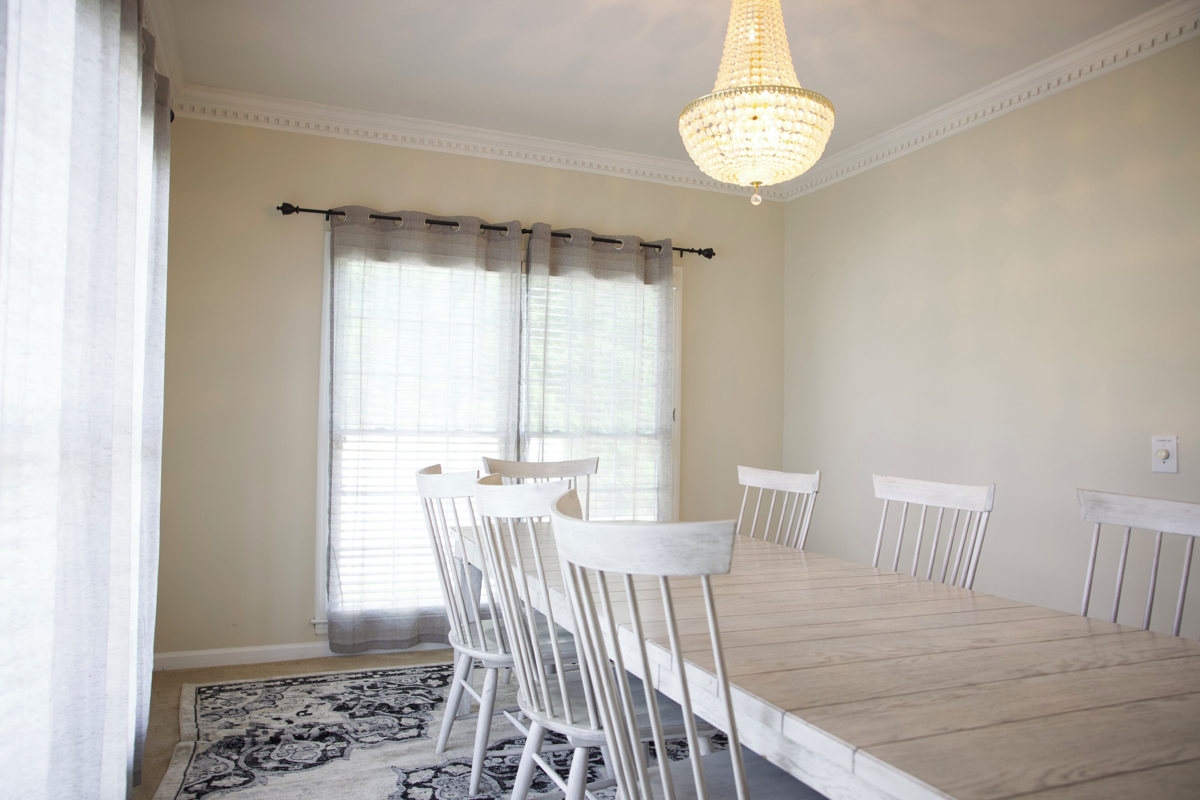 A dining room with a long table and white chairs, a chandelier, and a patterned rug.