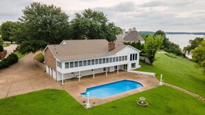 Une grande maison avec une piscine, entourée de pelouses verdoyantes et d’arbres, et offrant une vue sur un plan d’eau.