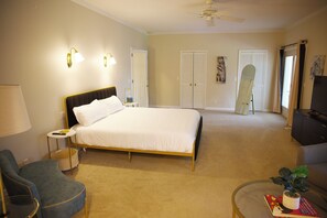 Quarto de hotel com cama, cadeira, pequena mesa com abajur, televisão e ventoinha no teto.