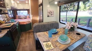 A compact RV interior with a kitchenette, a dining area with a table set for two, and a window providing a view of the outdoors. - Cozy riverfront RV in the heart of wine country "Dandy @ Verde Retreat" (Cottonwood)