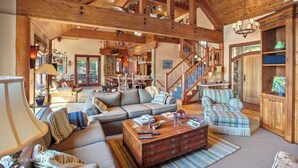 A spacious living room with a high ceiling, wooden beams, a staircase, and a cozy seating area with a coffee table. - Chateau Lac Hayden-Lake Front Luxury w/ Boats & Polaris rentals, lots of toys (Hayden)