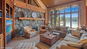 A cozy living room with stone walls, wooden beams, and large windows overlooking a lake. - Chateau Lac Hayden-Lake Front Luxury w/ Boats & Polaris rentals, lots of toys (Hayden)