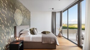 A modern bedroom with a large bed, a nightstand, a vase with dried flowers, and a view of the countryside through large windows. - Lovely home in Brtonigla with sauna (Brtonigla)