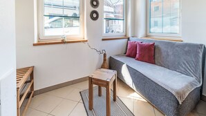 Un accogliente soggiorno con un divano grigio, un tavolino in legno e un tappeto decorativo. Sono presenti tre finestre con tende e un elemento decorativo sulla parete sopra il divano.