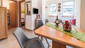 Zona pranzo con tavolo in legno, due sedie nere, tovaglia verde, una bottiglia di vino e un bicchiere. In parete, una televisione, uno specchio e una finestra con vista sugli edifici.