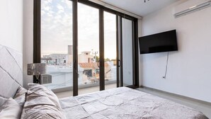 A bedroom with a large window, a bed, a flat-screen TV mounted on the wall, and a ceiling fan.