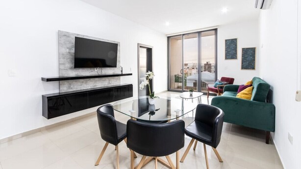 A modern living room with a glass dining table, black chairs, a green sofa, and a flat-screen TV mounted on a marble shelf.
