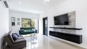 Ein modernes Wohnzimmer mit einem an der Wand montierten Flachbildfernseher, einem grauen Sofa, einem kleinen Couchtisch und einem Fenster mit Blick auf Grünflächen.